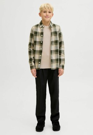 Camisa de franela a cuadros en verde, beige y verde oscuro con botones, usada sobre un suéter beige de canalé, combinada con pantalones negros y zapatos oscuros.