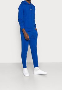 Persoon draagt een blauwe Calvin Klein hoodie en bijpassende joggingsbroek met witte sneakers, staand tegen een effen witte achtergrond.