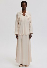 Ljust beige blus med stora fickor och krage, ihop med en matchande lång kjol. Slät textur, minimalistisk design och skräddarsydd passform.