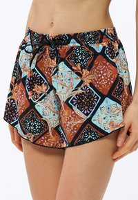Κοντινή λήψη ατόμου που φοράει πολύχρωμα σορτς με κορδόνι στη μέση και σχέδια με λουλούδια και γεωμετρικά σχήματα σε ανοιχτό φόντο.