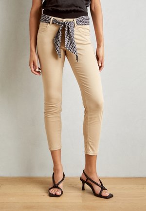 Pantalon skinny beige avec une ceinture à nouer noire et blanche, présentant une texture lisse et une longueur à la cheville fuselée. Porté avec des sandales à talons.