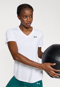 Baltas trumpomis rankovėmis marškinėlis su V formos iškirpte ir juodu logotipu, derinamas su žalsvais tamprėmis. Asmuo laiko juodą medicininį kamuolį.