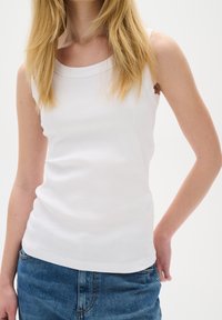 Femme blonde portant un débardeur blanc sans manches à côtes et un jean bleu, debout devant un fond uni.