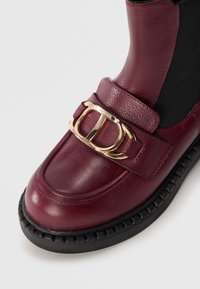 Mocassino in pelle bordeaux con dettagli in oro e suola nera in gomma spessa su sfondo bianco.