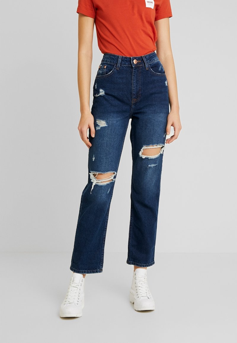 Höga, mörkblå denimjeans med slitna knäskärningar, rak benstil och kontrasterande sömmar. Kombinerat med vita sneakers.