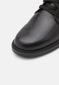 Zapato de cuero negro con punta redondeada, textura suave, diseño de cordones y suela de goma de contraste. Costuras visibles a lo largo del borde.