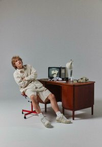 Tenue rayée beige clair, bureau en bois avec lampe rétro, téléphone vintage et vieille télévision affichant des notes autocollantes, avec une chaise rouge à roulettes.