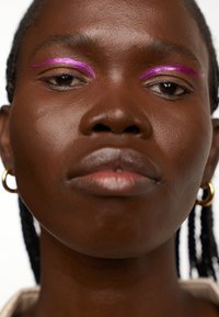 Gros plan sur un visage à la peau foncée, portant des boucles d'oreilles en or, et un eye-liner rose vif au-dessus des yeux, mettant en valeur une texture lisse et des lèvres subtiles.