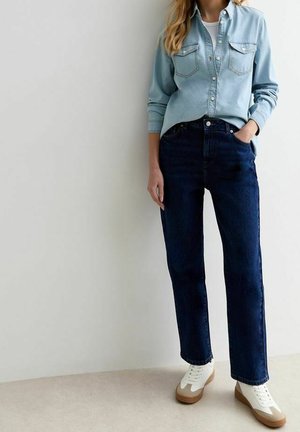 Henkilö, jolla on päällään vaaleansininen farkkupaita, tummansiniset farkut ja valkoiset tennarit, joissa on beige pohja, seisoo valkoista seinää vasten.