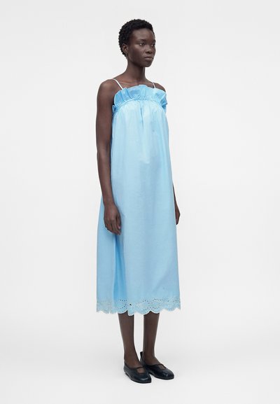 Γυναίκα με ανοιχτό μπλε μίντι αμάνικο φόρεμα με βολάν στο πάνω μέρος και σκίσιμο στο τελείωμα, σε συνδυασμό με μαύρα επίπεδα παπούτσια, όρθια μπροστά σε μονόχρωμο φόντο.