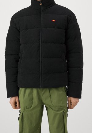 Personne portant une veste matelassée noire avec un petit logo coloré sur la poitrine et un pantalon cargo vert avec des poches zippées.