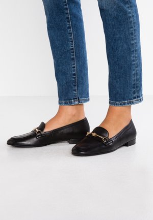 Czarne skórzane mokasyny na płaskiej podeszwie, z niewielkim złotym ozdobnym elementem i zaokrąglonym noskiem. Noszone z dopasowanymi niebieskimi jeansami.
