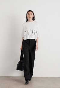 White cropped t-shirt with "CALVIN" printed in black, paired with black wide-leg trousers and a black bag. Model showcases a minimalist look.