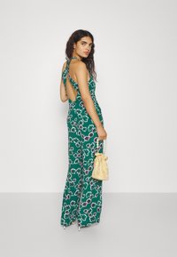 Grønn blomstrete jumpsuit med åpen rygg, vide ben og tynne stropper; sammen med en teksturert gul veske og høye hæler.