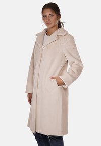 Cappotto in pelliccia sintetica color beige chiaro con colletto a scollo, maniche lunghe, tasche frontali e design aperto davanti. Texture morbida con una finitura liscia.