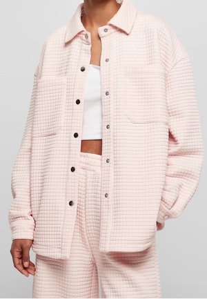 Person trägt hellrosa gesteppte Jacke mit Knöpfen und passende Hose über einem weißen Cropped Top, dargestellt vom Hals bis zur Mitte des Oberschenkels.