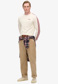 Beige overhemd met lange mouwen en rode zijstrepen, geruite flanellen stof om de taille, tan corduroypantalon en lichte sneakers met een gestructureerd oppervlak.