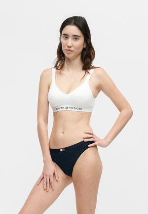 Νεαρή γυναίκα με λευκό bralette Tommy Hilfiger και σκούρο μπλε εσώρουχο στέκεται μπροστά σε απλό ανοιχτό φόντο με το ένα χέρι στη μέση.