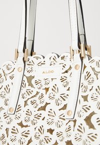Bolso tote blanco con diseños de mariposas recortados. Acabados en dorado, asas de hombro negras. Textura suave con una apariencia distintiva en capas.
