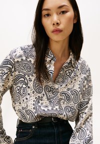 Vrouw met lang donker haar in een witte blouse met paisleypatroon, ingestopt in donkere spijkerbroek met een zwarte riem, tegen een effen achtergrond.