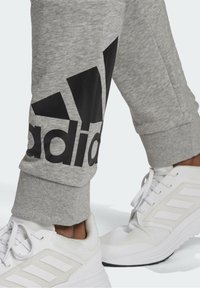 Grå sweatpants ben med stort sort Adidas-logo over det ribbede manschet, parret med hvide Adidas sportssko på lys baggrund.