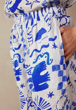 Witte linnen broek met een elastische tailleband, versierd met blauwe abstracte dierlijke en geometrische prints, met een hand in de zak.