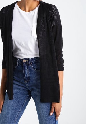 Donna che indossa una maglietta bianca, un cardigan nero a maglia aperta con motivi geometrici e jeans blu a vita alta su uno sfondo neutro.