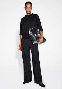 Haut en tricot noir avec un col montant, associé à un pantalon large noir. Le modèle tient un sac fourre-tout métallisé argenté. Chaussures noires avec de petites accents.