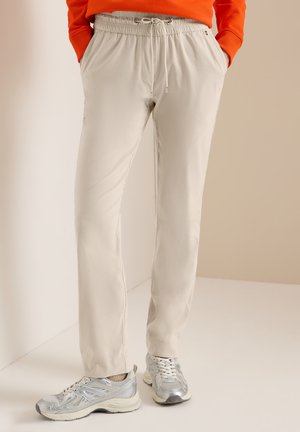 Personne portant un pantalon beige à cordon, un haut orange et des baskets argentées, debout les mains dans les poches sur un fond neutre.