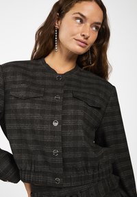 Vrouw met lang bruin haar draagt een zwart geruite geknoopte cropped jack en hangende kristallen oorbellen, poserend tegen een witte achtergrond.