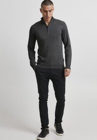 Tamno sivi pleteni pulover s polu-zip ovratnikom, s rebrastim manžetama i rubom, u kombinaciji s crnim suženim hlačama i crnim tenisicama.