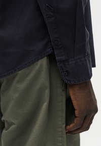 Χέρι με σκουρόχρωμο δέρμα που ξεκουράζεται στο πλάι, φοράει σκούρο μπλε πουκάμισο με μακριά μανίκια και παντελόνι χακί, εμφανίζεται από τη μέση έως τον καρπό.