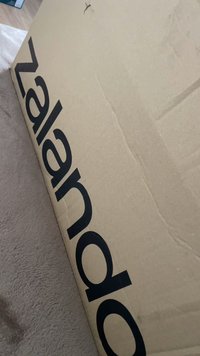 Barna kartondoboz, oldalán nagy fekete "Zalando" logóval, melyen enyhe kopás és egy kis szakadás látható.