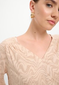 Blusa beige con motivo, scollatura a V e maniche arricciate. Tessuto liscio con disegni marroni ondulati. Modella che indossa orecchini a cerchio dorati.