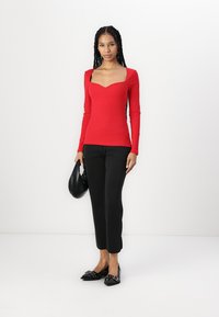 Rød ribbet langermet topp med firkantet hals, paired med svarte cropped bukser og svarte loafers. Modellen holder en svart håndveske.