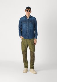 Denim ing két mellzsebbel, olajzöld cargo nadrág több zsebbel, és bézs sportcipő. A modellen napszemüveg és sapka van.