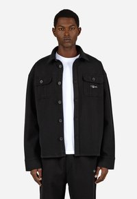 Fekete gombos kabát, textúrált anyagból készült, két mellzsebbel, gallérral és alatta egy fehér pólóval.