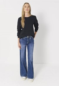 Eine Frau steht vor einem weißen Hintergrund und trägt ein schwarzes Langarmoberteil, blaue Schlagjeans und weiße Sneakers, die Hände entspannt an den Seiten.