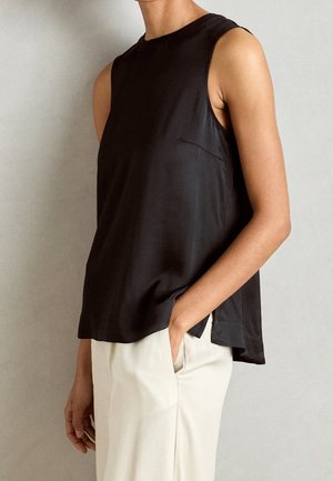 Top negro sin mangas de tejido liso con escote redondo; presenta una abertura lateral y un pequeño bolsillo en el pecho, combinado con pantalones de color claro.