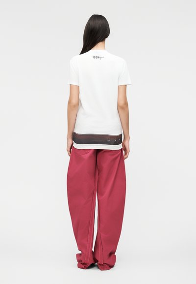 Γυναίκα με μακριά σκούρα μαλλιά φοράει λευκό κοντομάνικο πουκάμισο με μαύρη ρίγα κοντά στη ραφή και φαρδιά κόκκινα παντελόνια, στέκεται με την πλάτη γυρισμένη.