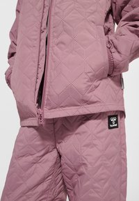 Pikowana fioletowa kurtka i spodnie z geometrycznym przeszyciem, ręce w kieszeniach kurtki, widoczne logo hummel na spodniach.