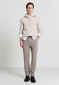 Maglione beige lavorato a maglia con collo alto e mezza cerniera, indossato sopra una camicia bianca. Pantaloni sartoriali marrone chiaro e scarpe nere completano il look.