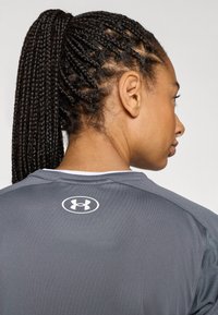 Szürke atlétikai póló rövid ujjakkal, hátul fehér logóval. Az anyag simának és kissé texturáltnak tűnik.