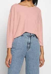 Hellrosa, gerippter Strickpullover mit weitem Ausschnitt und Drei-Viertel-Ärmeln, kombiniert mit hellblauen, hochgeschnittenen Jeans.