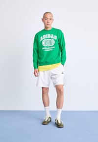 Grön sweatshirt med vit logotyp, lager på lager över en gul skjorta, vita shorts med svarta accenter och gröna sneakers med vita ränder.
