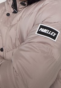 Puchowa, beżowa kurtka z gładkiego materiału. Posiada czarną naszywkę z napisem "PARELLEX" w białym kolorze na lewym rękawie. Widoczne guziki i kołnierz.