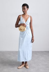 Ανοιχτό μπλε αμάνικο μάξι φόρεμα με βαθύ V-ντεκολτέ, υφαντό ύφασμα; σε συνδυασμό με μια υφασμάτινη τσάντα από ψάθα και μπεζ φλατ παπούτσια.