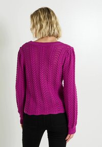 Pull en maille magenta avec un motif en zigzag et des poignets côtelés. Présente une longueur légèrement raccourcie et une texture en tissage ouvert.