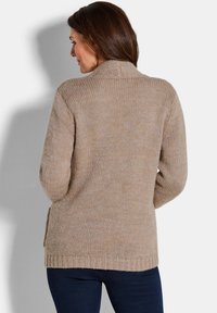 Frau mit braunen Haaren, die einen beigen Strickpullover und dunkle Hosen trägt, von hinten vor einem schlichten hellen Hintergrund gezeigt.