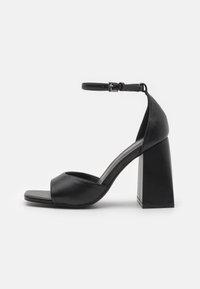 Des sandales à talons noirs présentent un dessus en cuir lisse, un bout ouvert, une sangle de cheville avec boucle et un large talon carré.